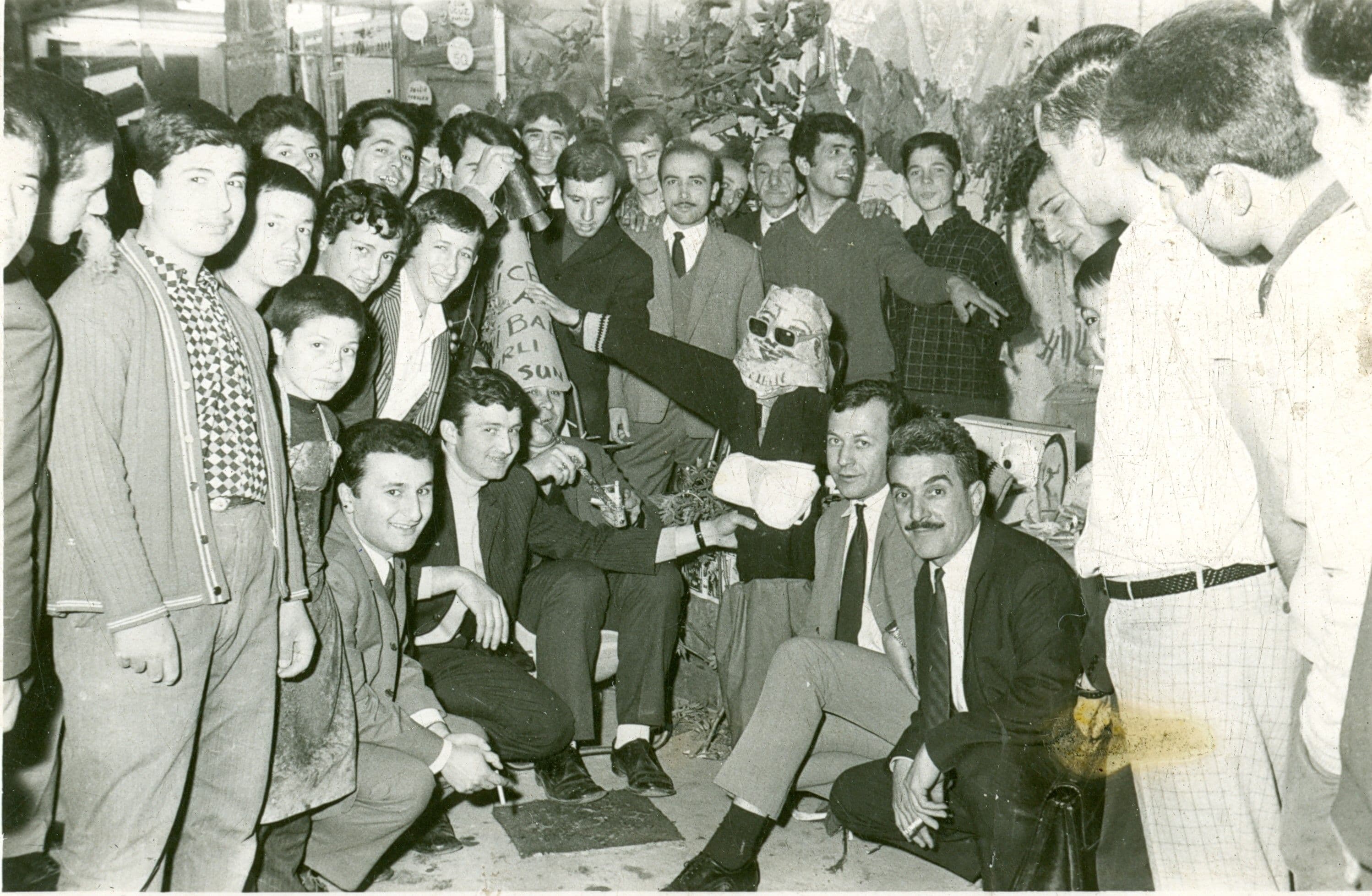 Eski çarşıda Hıdırellez kutlaması yapan bir grup esnafın siyah beyaz fotoğrafı; ortadaki bir kişi komik bir maske takıyor.