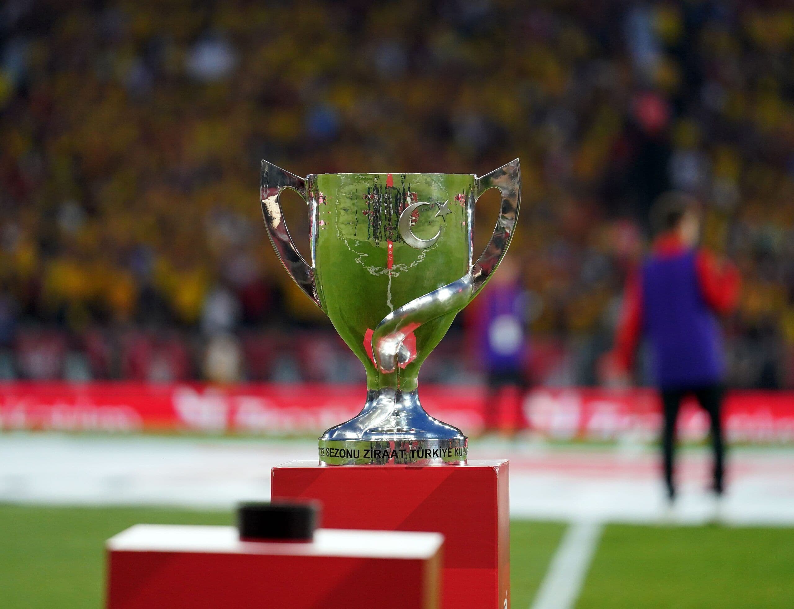 Ziraat Türkiye Kupası kupası, bir stadyumun zemininde kaide üzerinde duruyor, arka planda bulanık tribünler görünüyor.