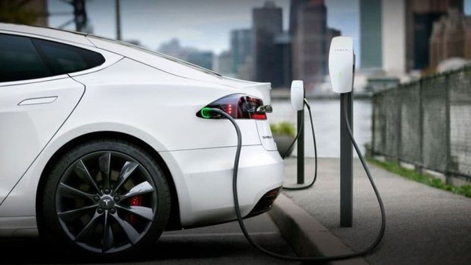 Beyaz bir elektrikli araç şarj istasyonunda şarj oluyor, şehir manzarası arka planda.