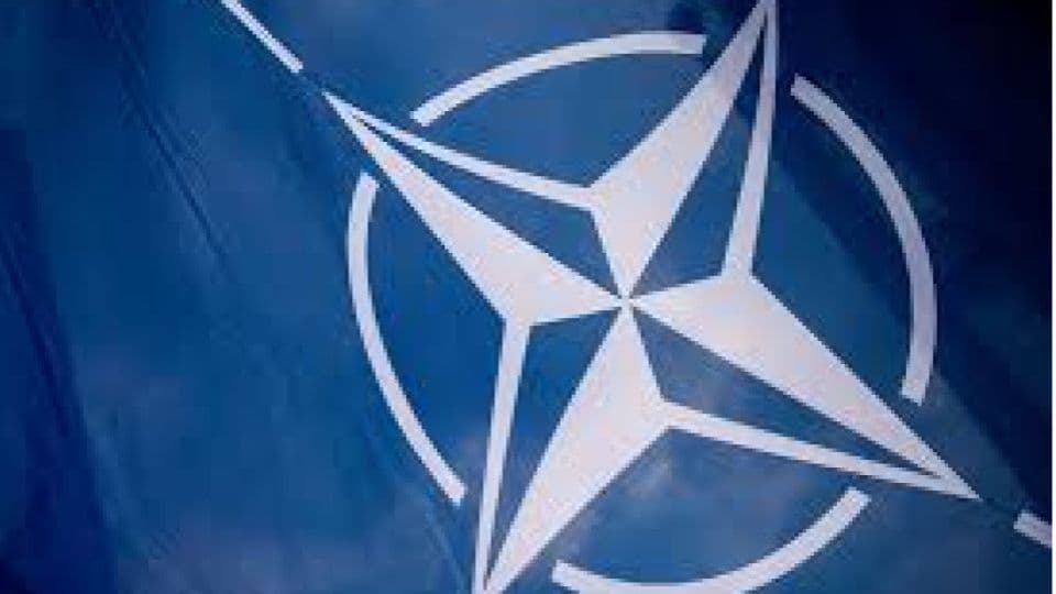 Mavi zemin üzerine beyaz pusula gülü sembolü olan dalgalanan NATO bayrağı.