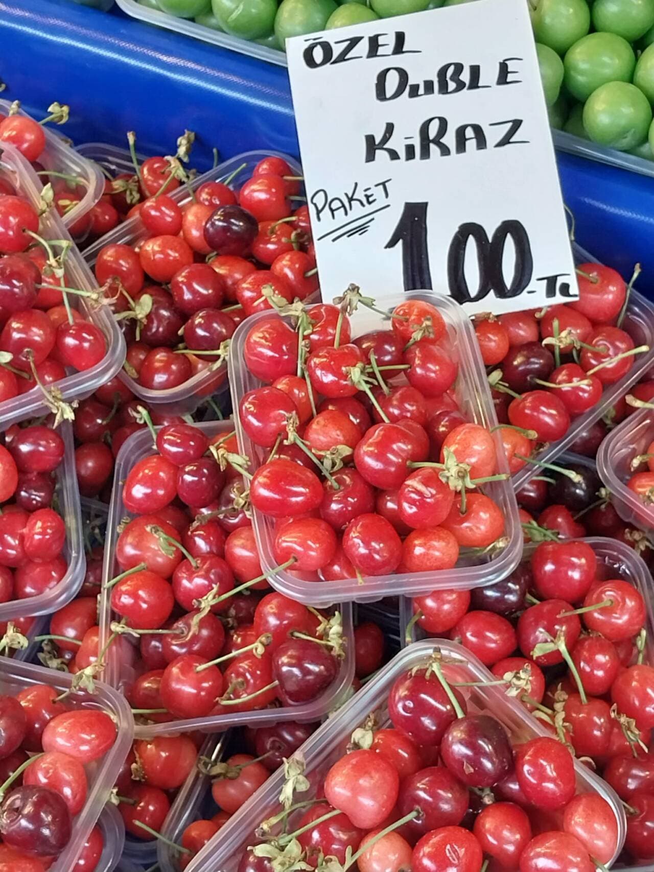 Pazar tezgahında plastik kaplar içinde sergilenen taze kırmızı kirazlar ve üzerinde 'Özel Duble Kiraz Paket 100 TL' yazan bir fiyat etiketi.