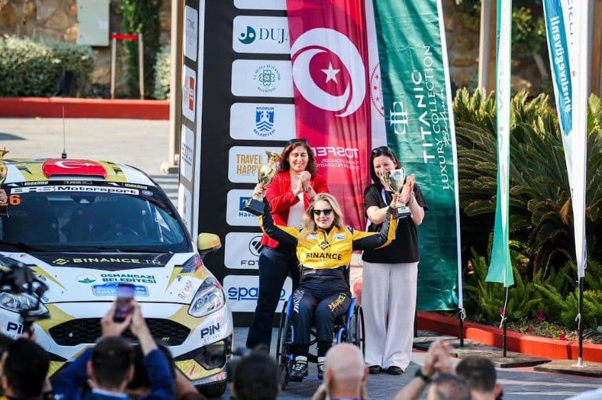 Bursalı pilot Kübra Denizci Keskin, Bodrum Rallisi'nde kazandığı kupalarla podyumda, yanında ralli aracı.