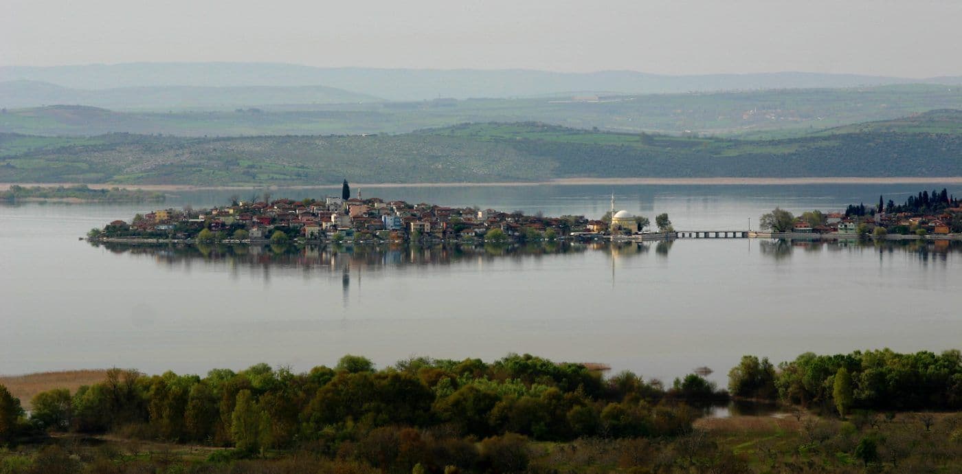 Uluabat Gölü'ndeki Gölyazı köyü, evleri, camisi ve köprüsüyle bir adada yer alıyor. Arka planda yeşil tepeler ve göl manzarası.