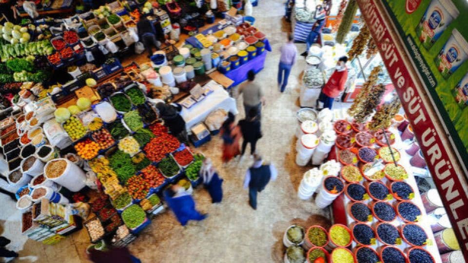 Kalabalık bir pazar yerinde sebze, meyve ve baharat tezgahları.