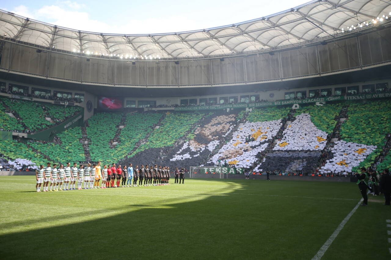 Bursaspor stadyumunda maç öncesi seremoni, futbolcular sahada dizilmiş, tribünlerde yeşil-beyazlı taraftarların koreografisi.