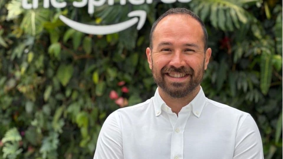 Amazon yöneticisi Yağız Ihlamur gülümsüyor, arkasında Amazon logosu ve yeşil bitkiler var.
