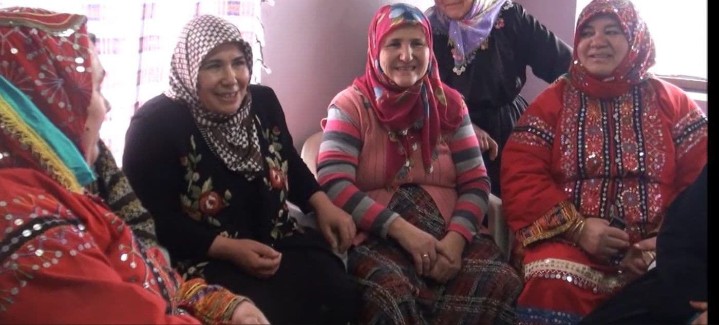 Geleneksel kıyafetler giymiş, başörtülü, gülümseyen yaşlı kadınlar bir araya gelmiş, mânilerin yaşatıldığı bir ortamda sohbet ediyor.