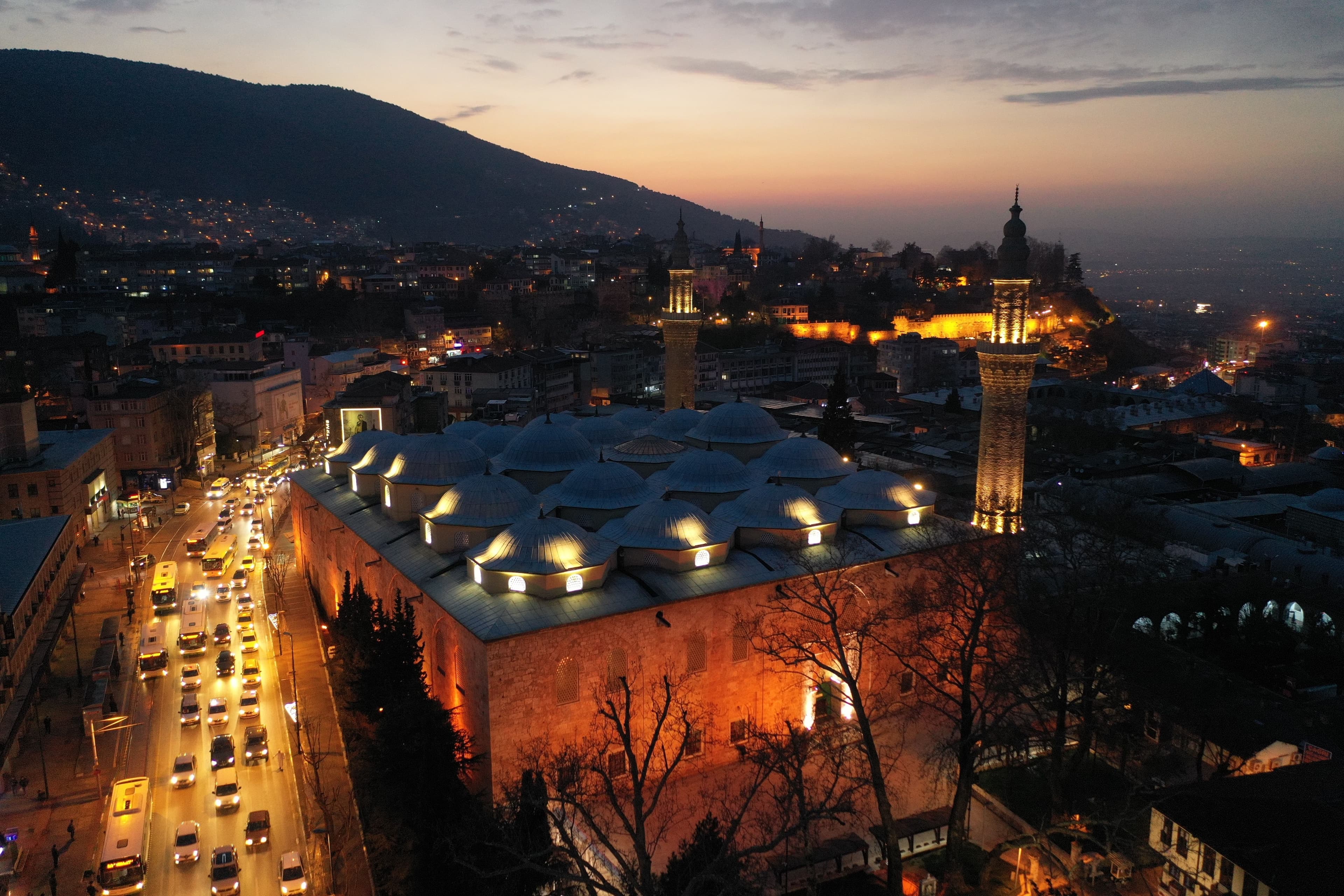 Gün batımında ışıklandırılmış Bursa Ulu Cami'nin, çevresindeki şehir ve dağ manzarasıyla birlikte çekilmiş havadan fotoğrafı.