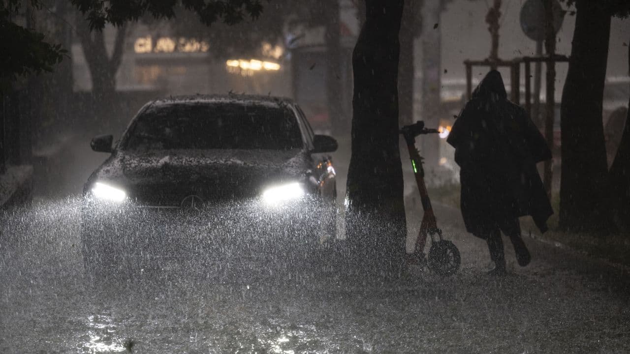 Gece sağanak yağış altında farları açık bir araba ve kaldırımda yürüyen yağmurluklu bir kişi.
