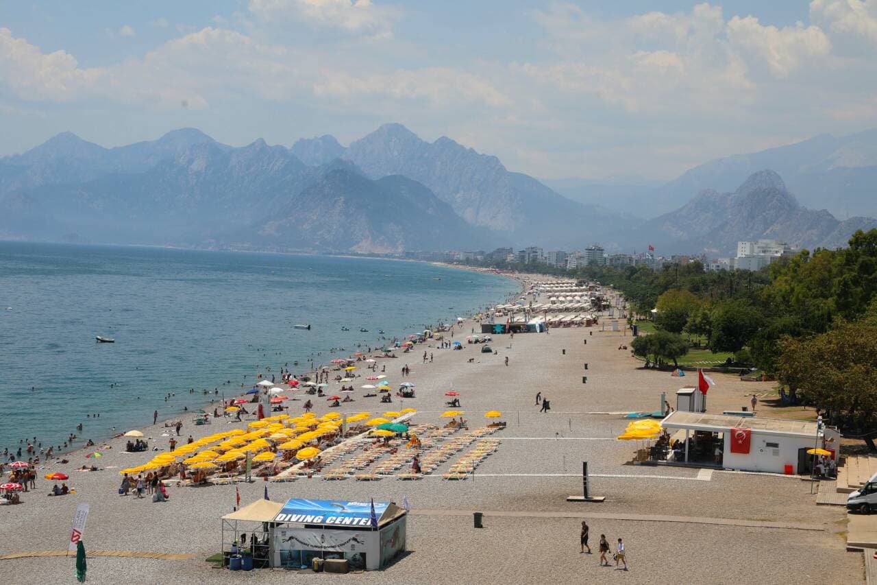 Antalya Konyaaltı Sahili'nde sıcak havada denize giren ve güneşlenen insanlar, kalabalık plaj ve dağ manzarası.