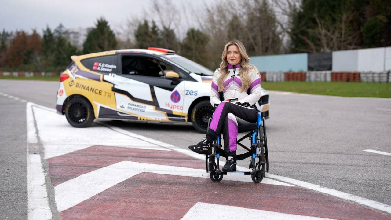 Engelli ralli pilotu Kübra Denizci Keskin, yarış kıyafetleriyle tekerlekli sandalyesinde otururken, arkasında ralli aracıyla birlikte pistte görülüyor.