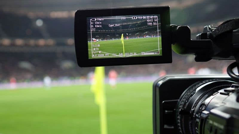 Bir futbol maçını canlı yayınlayan televizyon kamerasının ekran görüntüsü, sahada oynayan futbolcular ve tribünler.