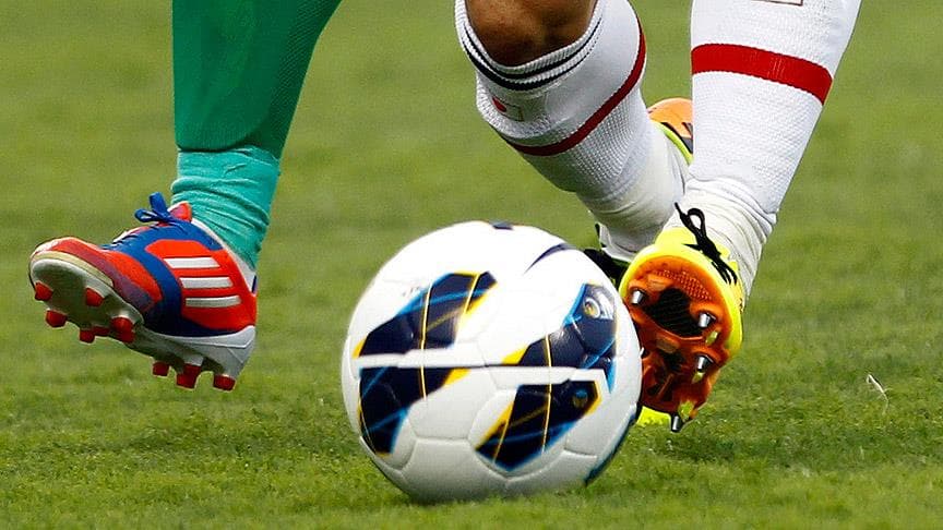 Futbol topu ve futbolcuların kramponlu ayakları, yeşil sahada maç anı.