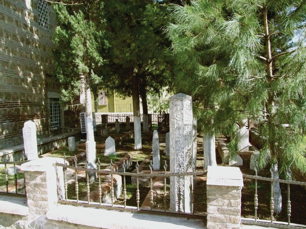 Bursa'daki tarihi bir hazireden görünüm. Farklı dönemlere ait Osmanlı mezar taşları, yeşil ağaçlar ve taş bir yapının yanında yer alıyor.