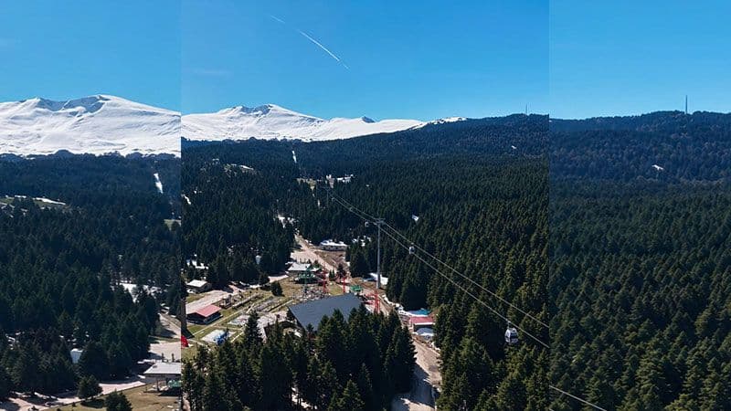 Uludağ'ın zirvesinde kar, yamaçlarında bahar yeşillikleri ve ağaçlar, teleferik hattı.
