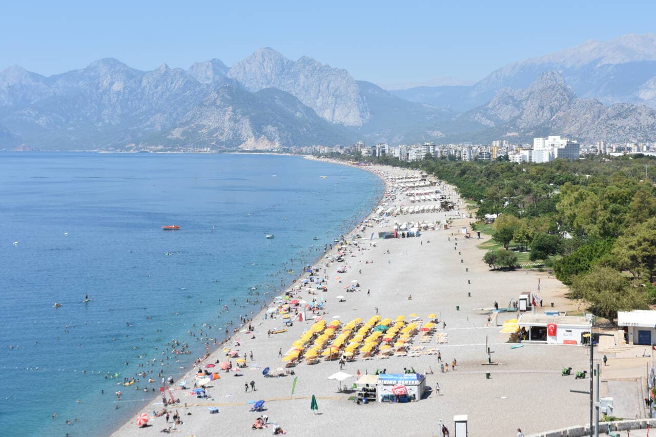Antalya Konyaaltı veya Lara sahilinde sıcak havada denize giren ve güneşlenen kalabalık insanlar, dağ manzarası.